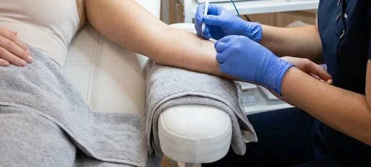 A gloved practitioner guiding a fine electrolysis probe near a reclining client’s arm beside a magnifying lamp and control unit.